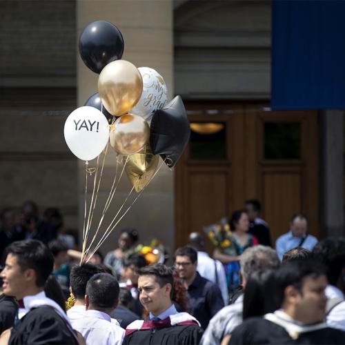 Balloons with the words "YAY!" and "Congrats Grads" catch a shaft of sunlight above a crowd outside Convocation Hall.