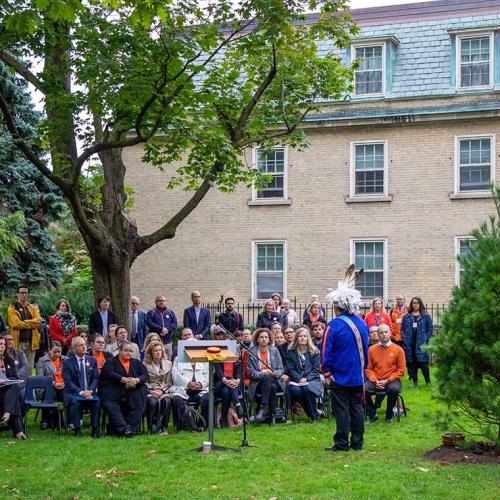 Skahendowaneh Swamp speaks to a crowd gathered on a green lawn. Behind him is a newly planted Eastern white pine tree.