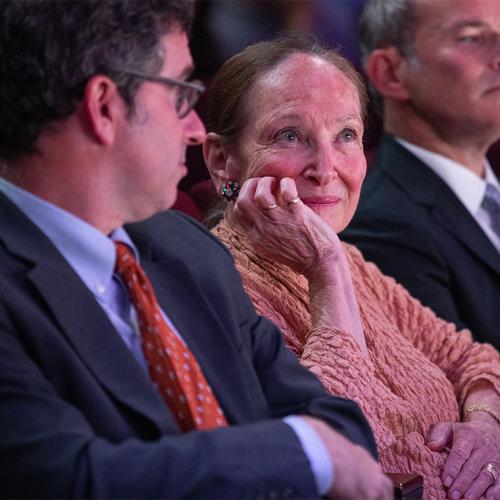 Rosie Abella sitting in an audience, listening, smiling, and resting her chin on one hand.