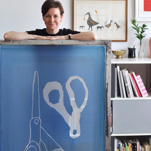 Alanna Cavanagh standing behind a desk in front of her artwork. 