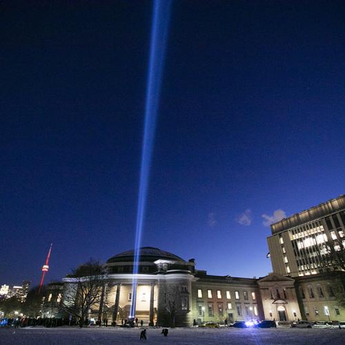 A spotlight shines straight up into the night sky on a snow-covered Front Campus, in front of a lit-up Convocation Hall.