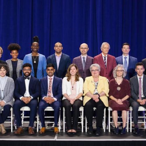 17 winners of Awards of Excellence pose on a stage, standing in two rows for a group picture and smiling. (photo by Gustavo Toledo Photography)