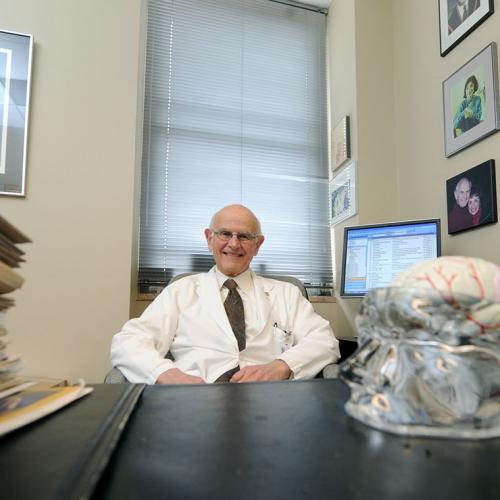 Charles Tator grins as he sits at his office desk, which holds a plastic model of the human brain.