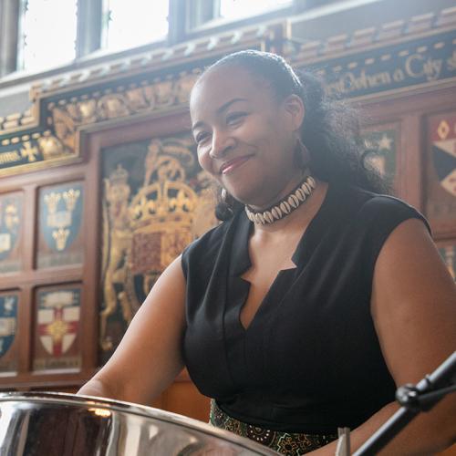Suzette Vidale smiles happily as she plays a drum in front of a panelled wooden wall at Hart House.