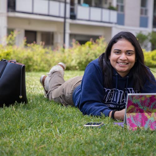 Amanda Khan smiles as she looks up from her laptop while lying on her stomach on a grass lawn.