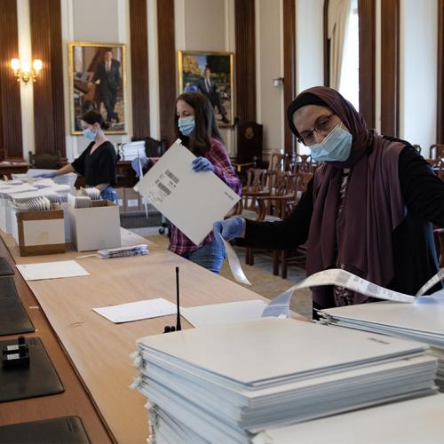 Three women wearing masks and gloves work at a long table loaded with boxes, lists, and piles of envelopes.