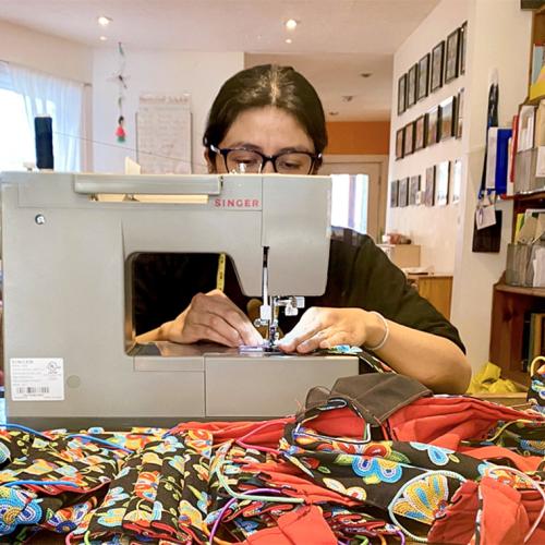 Fernanda Yanchapaxi sits behind a sewing machine; the table in front is covered with homemade cloth masks.