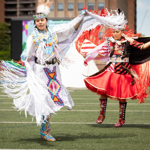 Alanna Pasche and Deanne Hupfield, wearing Indigenous regalia with fringe and embroidery, dance on the turf at Varsity Stadium.