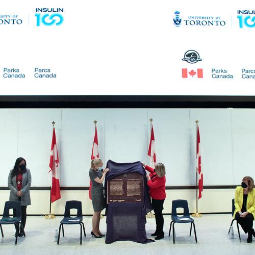 At a podium in a classroom, Christine Allen and Christine Loth-Brown lift a cloth to unveil a metal plaque.