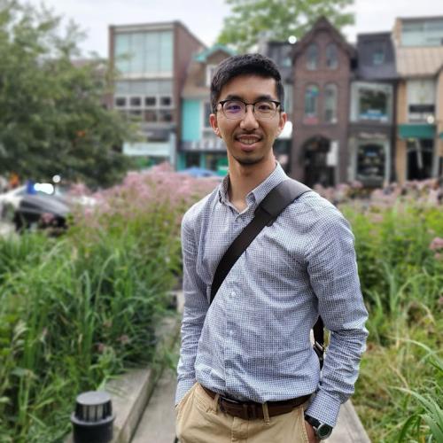 Lyndon Chan smiles while standing in a park beside a shopping street.