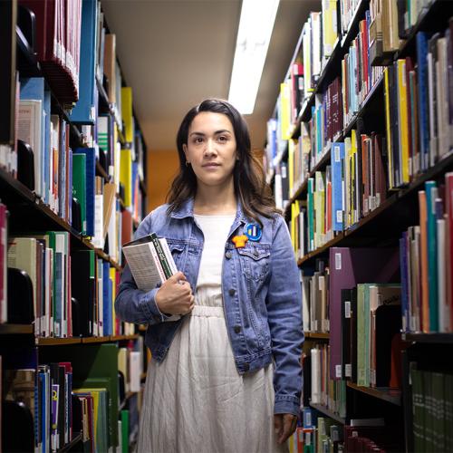 Mikayla Redden stands between tall library shelves, holding a stack of books and looking doubtful.