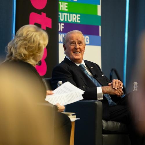 Brian Mulroney smiles as he sits in a chair on stage chatting with a moderator.