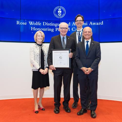 Paul Cadario stands on stage between Rose Patten, Corwin Cambray and Meric Gertler, holding his award and smiling