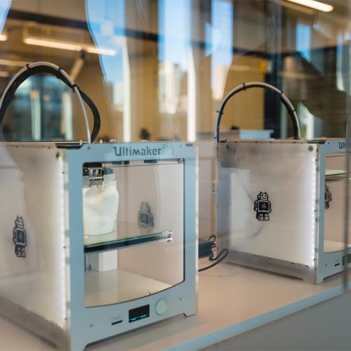 A row of 3D printers: plastic and metal boxes with a transparent front and a movable shelf inside.