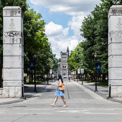 A woman in a mask walks past the U of T gateway on College Street, as the road stretches up to University College behind her.