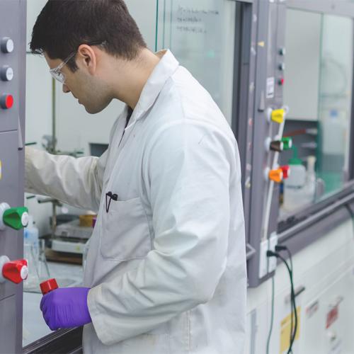 Wearing glasses and gloves, Aaron Cabal reaches into protectively walled lab bench that contains chemistry equipment.