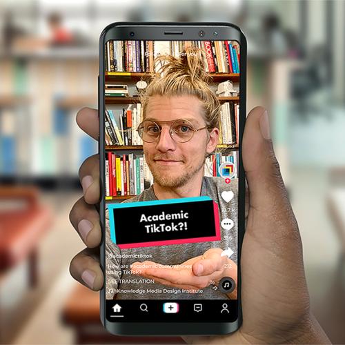 A hand holds a smartphone. On the screen, a young man makes a wry face. A text caption reads: Academic TikTok?!