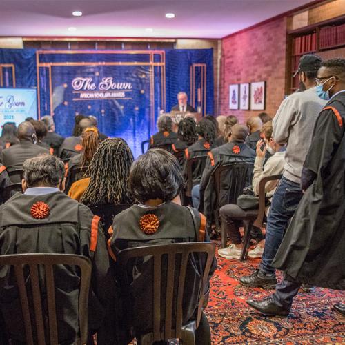 A group of people in masks sit in rows in a library room. They wear academic gowns with orange decorations.