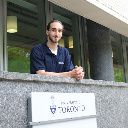 Aidan Gomez smiles, leaning on a stone balustrade above a sign that reads: University of Toronto.