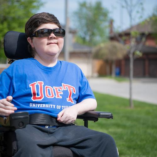 Alex Harold smiles and looks up as he sits outdoors with one hand on his wheelchair controls.