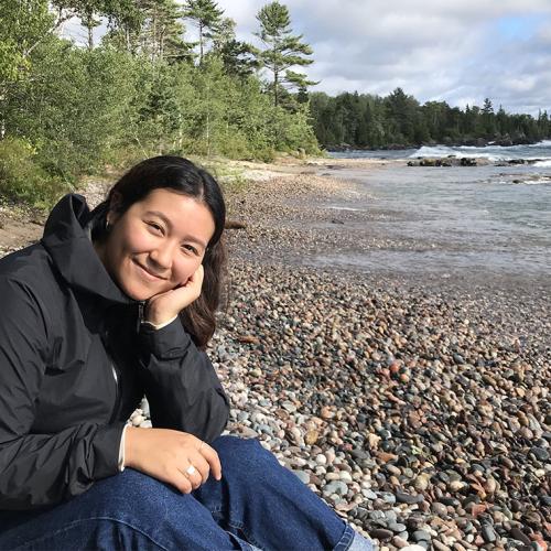 Alexandra Gaspar sits on a pebbled, tree-lined beach, leaning her head on one hand and smiling. Waves roll in behind her.