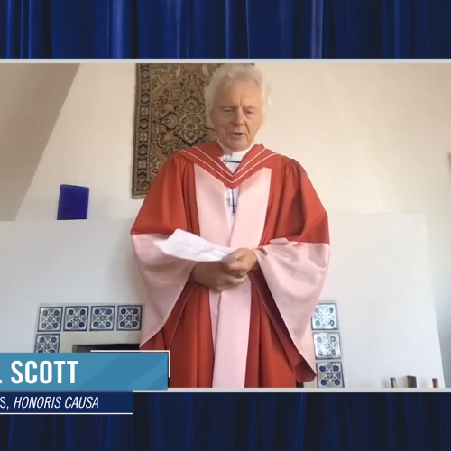 Allen J Scott, wearing academic robes, speaks from written notes while standing in front of a tiled fireplace.