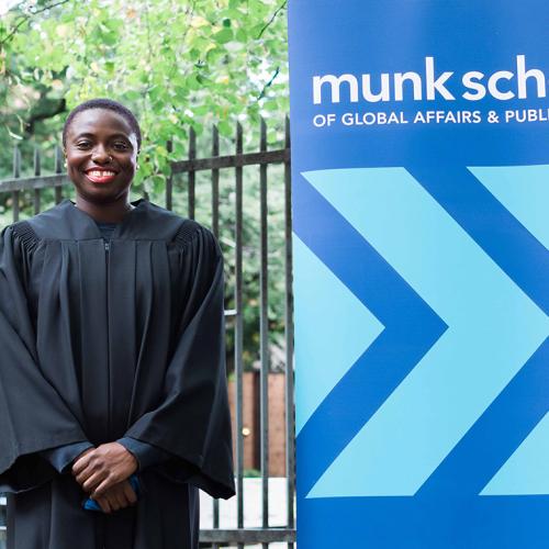 Anowa Quarcoo smiles happily. She is wearing academic robes and standing next to a sign for the Munk School.