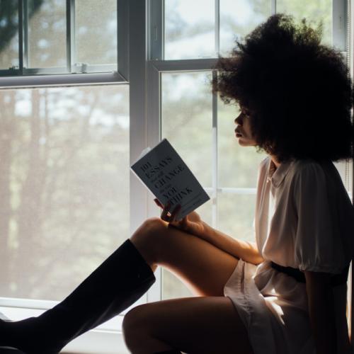 A young woman sits on a sunny windowsill, reading a book.