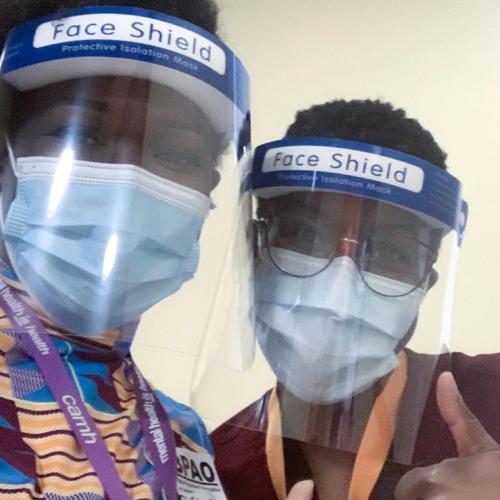 Onye Nnorom and Duate Adegbite, wearing masks and face shields, lean together and give the thumbs up.