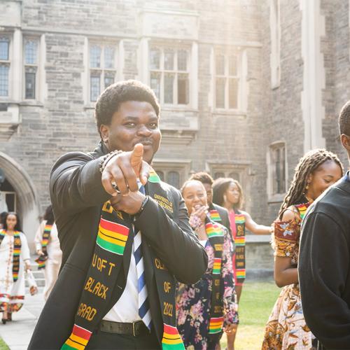 A young man smiles and points in celebration. He is wearing a stole with text on it: U of T Black Grad.