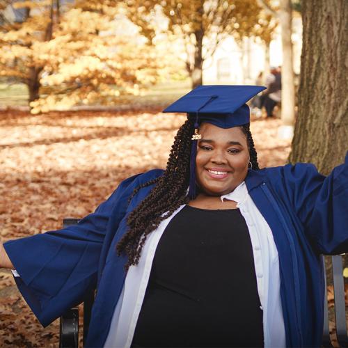 Carisse Samuel stretches out her arms and smiles, as she poses in academic cap and gown on a park bench.