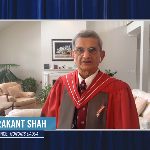 Chandrakant Shah, wearing academic robes, speaks on video from a living room with large plants.