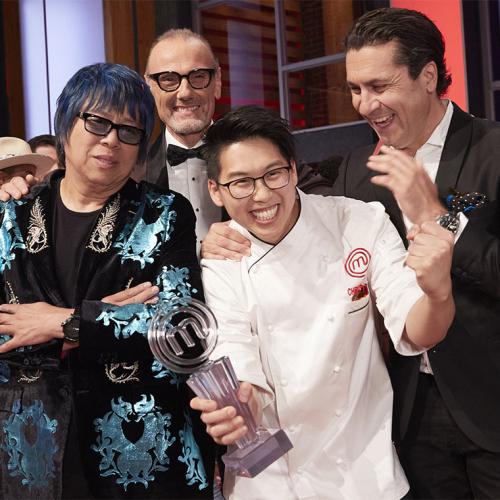Christopher Siu laughs and holds up a plexiglass trophy shaped like the letter M while three men smile and hug him.