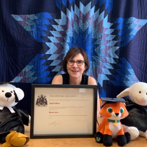 Cindy Blackstock smiles  in front of a quilt with a star pattern. On her desk are stuffed animals in academic robes and hats.