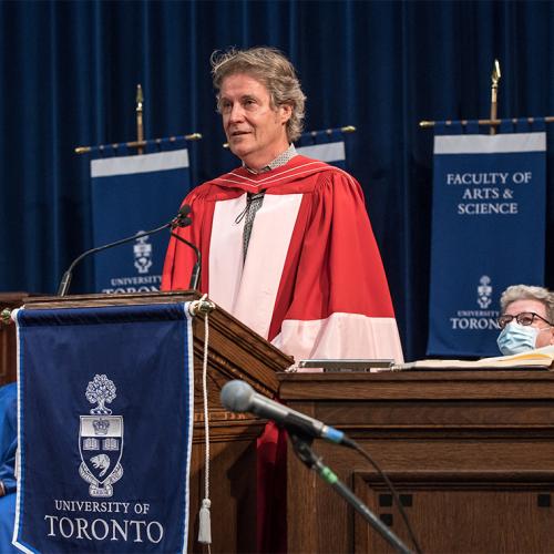 Jim Cuddy, wearing academic robes, speaks at the podium in Convocation Hall.