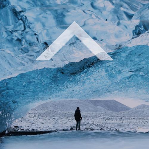 Man stands underneath an ice bridge looking at a mountain in the distance. A caret icon in the middle of the image.