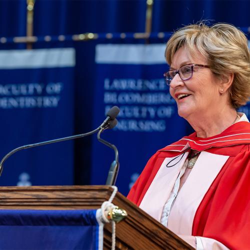Annette Kennedy smiles as she speaks at the podium in Convocation Hall, wearing academic robes.