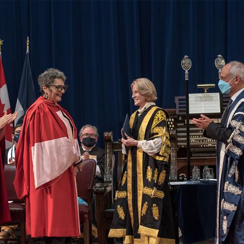 Nada Ristich smiles at Meric Gertler and Rose Patten, while a student puts on Nada's academic hood.