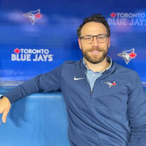 David Lawrence smiles, sitting in the Blue Jays dugout with the team logo behind him.