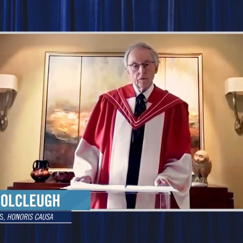 David Colcleugh, wearing his academic robes and hood, speaks on video in a room with an elaborate fireplace.