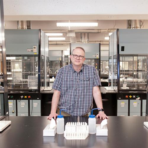David McMillen leans on a steel bench in a large lab full of equipment contained in glass and plastic hoods.
