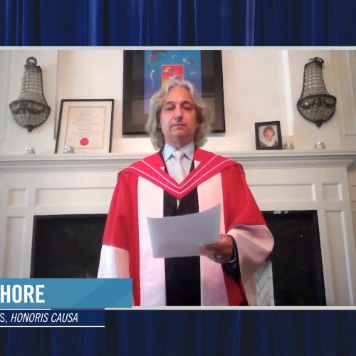 David Shore wears academic robes and speaks in front of a fireplace. On the mantel is his graduation picture as a young man.