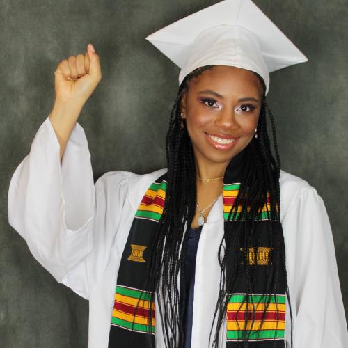 Denessia Blake-Hepburn raises her fist. She is wearing graduation robes and mortarboard hat, and a Black Grad kente scarf.
