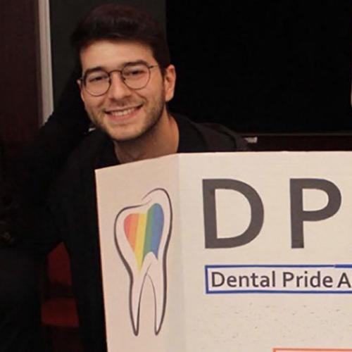 Lucia Santos and Evan Benak smile next to a poster with a rainbow-coloured tooth and the words: Dental Pride Alliance.