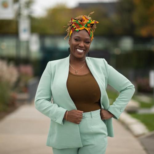 Desiree Kaunda-Wint  smiles, standing with hand on hip on a walkway outdoors.