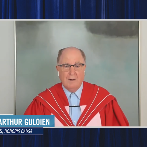 Donald Guloien, wearing academic robes, speaks while standing in front of an oil painting.