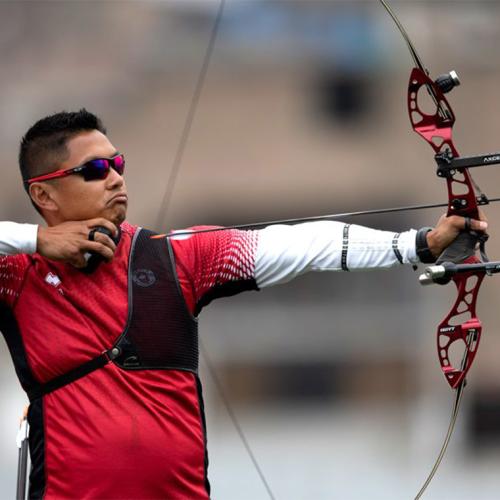 Crispin Duenas in the middle of loosing an arrow from his high-tech archery bow. He wears sunglasses and a chest protector.