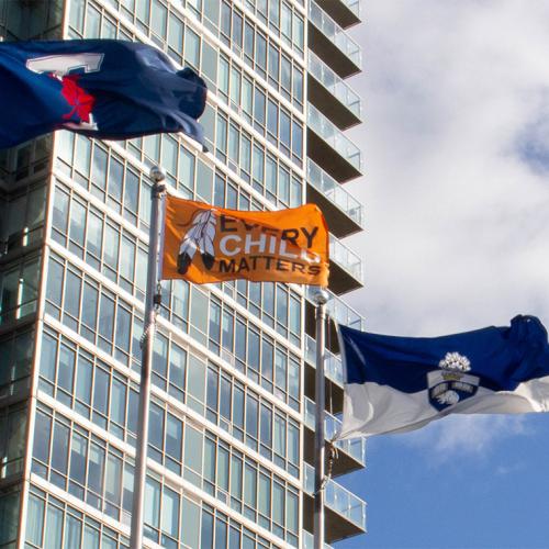 An orange flag with the words every child matters flies on a flagpole beside the U of T flag.