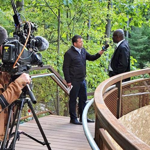 Wisdom Tettey chats with a CTV reporter on a curved wooden bridge, while a man points a large TV video camera at them.