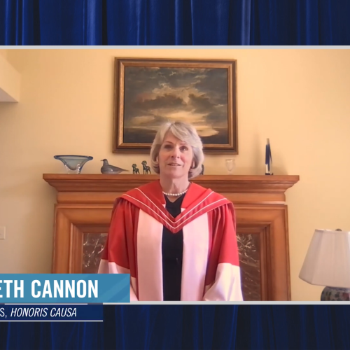 Elizabeth Cannon, wearing her academic robes and hood, speaks on video in a room with an elaborate fireplace.